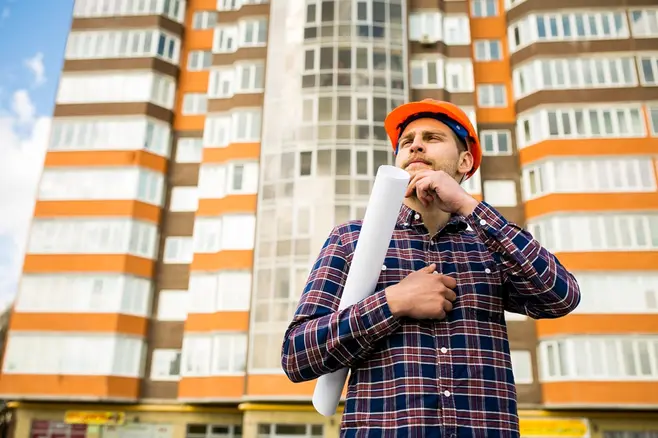 A man in a purple flannel shirt and a red construction helmet stands in front of an apartment building, holding a rolled-up sheet of paper under his arm